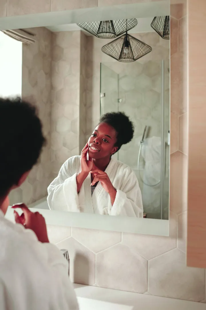 Woman in a soft-lit bathroom checking her skin in the mirror, warm natural light